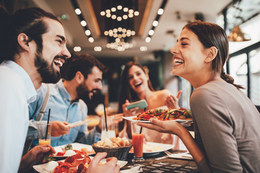 Group of happy people sitting at a restaurant table, smiling and laughing while enjoying their meal. The table is filled with delicious food and drinks, creating a warm and joyful atmosphere